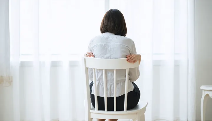 Pessoa sentada de costas em cadeira branca, mãos apoiadas nos joelhos, pés em movimento alternado, luz natural suave ao fundo