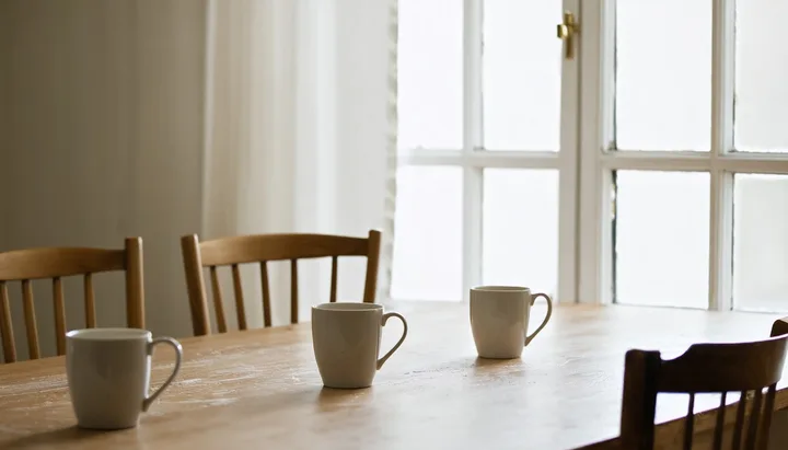 Mesa de cozinha com duas xícaras de café e cadeiras vazias, luz natural entrando pela janela, representando espaço de diálogo em família
